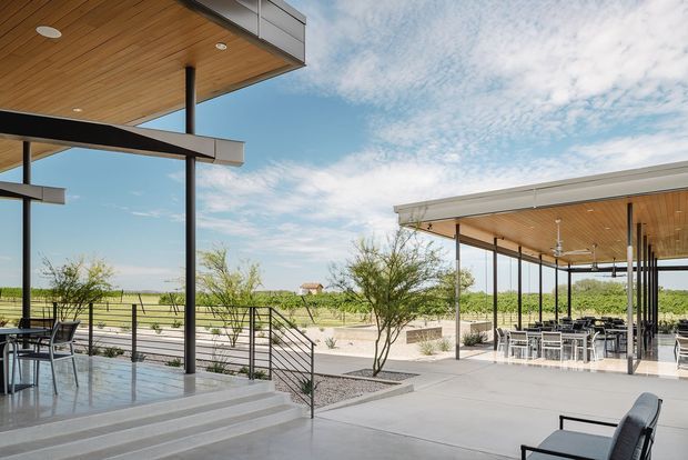 A contemporary outdoor hospitality or residential entertaining area with broad flat-roofed covered terraces, slim dark structural posts, warm timber soffits, and expansive views across an open rural landscape. The space includes a paved terrace, outdoor dining furniture, ceiling fans, steps, and minimalist metal balustrades, with landscaped gravel garden beds between the covered areas.