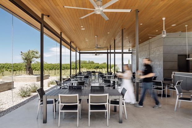 A contemporary covered dining terrace or hospitality venue with extensive floor-to-ceiling glazing, outdoor tables and chairs, a warm timber-lined ceiling, exposed dark metal columns, ceiling fans, and a polished concrete floor. A vineyard landscape is visible beyond the open glazed facade, while a dark concrete-clad service or bar wall defines the right side. The image has a bright, airy indoor-outdoor character.