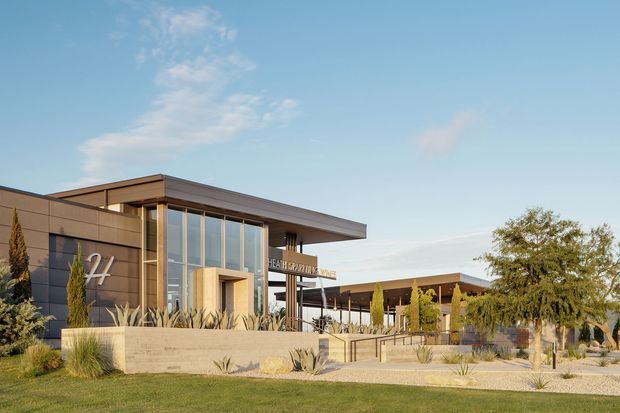 A low-rise modern commercial building viewed from the landscaped exterior. The facade combines dark horizontal cladding, extensive floor-to-ceiling glazing, a projecting flat roof, and a sheltered entrance reached by broad steps and accessible ramps. Drought-tolerant planting, sculptural grasses, and slender columnar trees surround the building. A covered outdoor walkway or pavilion extends to the right under a flat roof. The scene is photographed in clear daylight with a restrained contemporary architectural character.