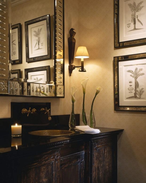 A richly styled, traditional bathroom vanity with a dark carved timber cabinet, polished black stone benchtop, and an inset oval basin with antique brass tapware. Warm textured beige walls are decorated with framed botanical prints and an ornate mirrored cabinet, while a wall sconce, candle, and tall glass vases create a moody, luxurious atmosphere.