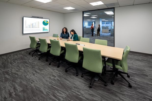 A modern commercial meeting room with a long light-wood conference table surrounded by green upholstered office chairs. The room has grey patterned carpet, white walls, a suspended acoustic tile ceiling with recessed lighting, and a wall-mounted presentation screen. A glazed partition provides views into adjacent office areas.