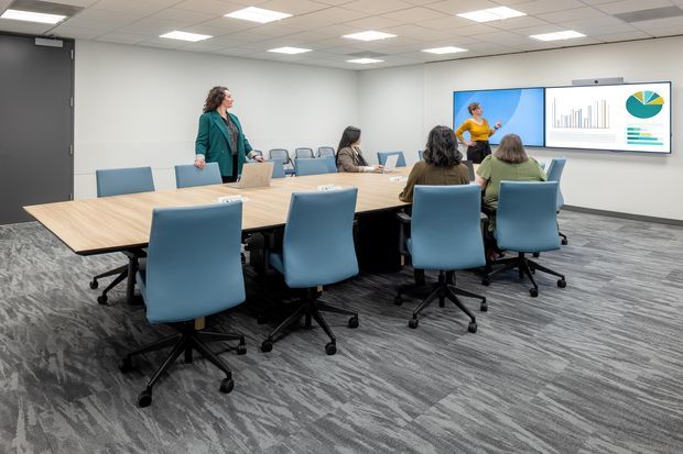 A modern corporate conference room with a large rectangular light-wood meeting table surrounded by blue upholstered swivel chairs. Several people are seated around the table while one person presents beside a wall-mounted display showing charts. The room has grey walls, a suspended acoustic tile ceiling with recessed lighting, and patterned grey carpet flooring.
