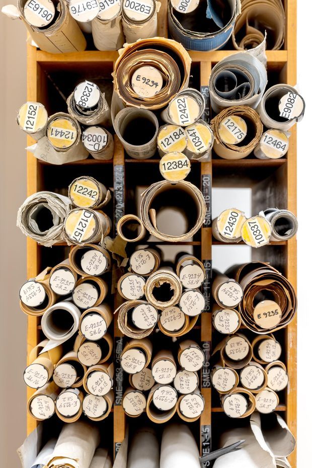 Top-down view of a timber storage rack filled with tightly rolled sheets or plans, each roll labelled with handwritten or printed identification numbers. The image is dominated by the organised shelving and cylindrical paper materials, with warm wood tones and neutral whites, greys, and browns.