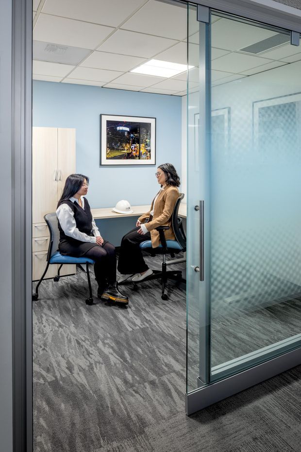 A small contemporary office meeting room viewed through a frameless glass door. Two people sit facing each other on office chairs beside a compact white table. The room has pale blue walls, light wood built-in storage, grey patterned carpet, a suspended acoustic tile ceiling with fluorescent lighting, framed artwork, and frosted patterned glass providing privacy. The image is a professionally styled commercial interior photograph.