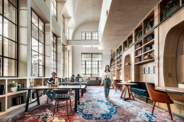 A bright, high-ceilinged contemporary communal workspace or library with expansive black-framed industrial-style glazing, exposed concrete surfaces, extensive built-in timber shelving and arched joinery, worktables, lounge seating, and a large patterned rug. Several people are using the desks, creating an active yet refined office or hospitality-like atmosphere.