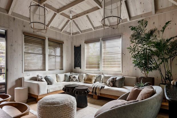 A warm, light-filled living room with a large L-shaped sectional sofa, additional curved lounge seating, a round woven ottoman, and a low dark coffee table. The room features pale timber wall panelling and exposed timber roof framing with a vaulted, geometric ceiling. Multiple windows have layered timber-toned blinds, while woven textiles, cushions, indoor plants, and black wire pendant lights add a relaxed natural character.