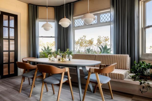 A bright, refined dining nook with a long upholstered banquette beneath large leaded-glass windows. A pale oval dining table is surrounded by mid-century-inspired timber and upholstered chairs, with three globe pendant lights overhead. Dark striped curtains, warm roller blinds, timber-look flooring, indoor plants, and a black-framed glazed door create a layered, elegant interior.