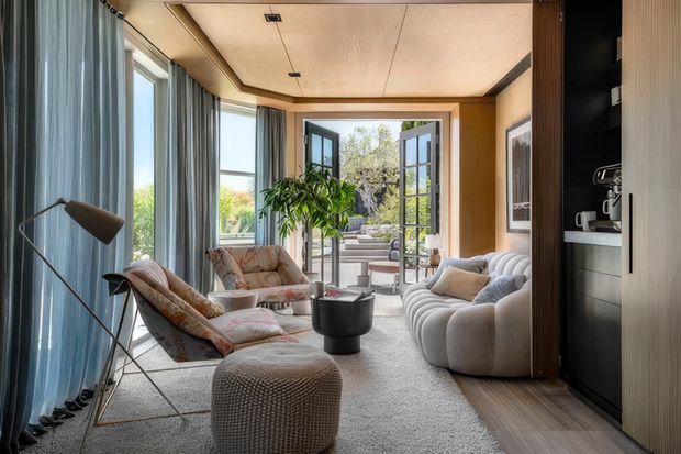 A bright, refined living room with floor-to-ceiling windows and sheer blue-grey curtains, a large plush white curved sofa, patterned lounge chairs, a textured round ottoman, and a black coffee table. Warm timber wall and ceiling finishes frame built-in shelving and a compact beverage station. Glazed doors open onto a landscaped outdoor area, bringing in abundant natural light.