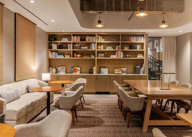 Warm, refined open-plan living and dining area with a full-height timber built-in shelving and cabinetry wall, upholstered seating, a round coffee table, and a large timber dining table. Recessed ceiling lights and pendant fixtures illuminate the room, while a dark exposed concrete ceiling beam adds contrast. Floor-to-ceiling curtains and blinds frame the windows, and a stairway is visible behind a glazed partition.