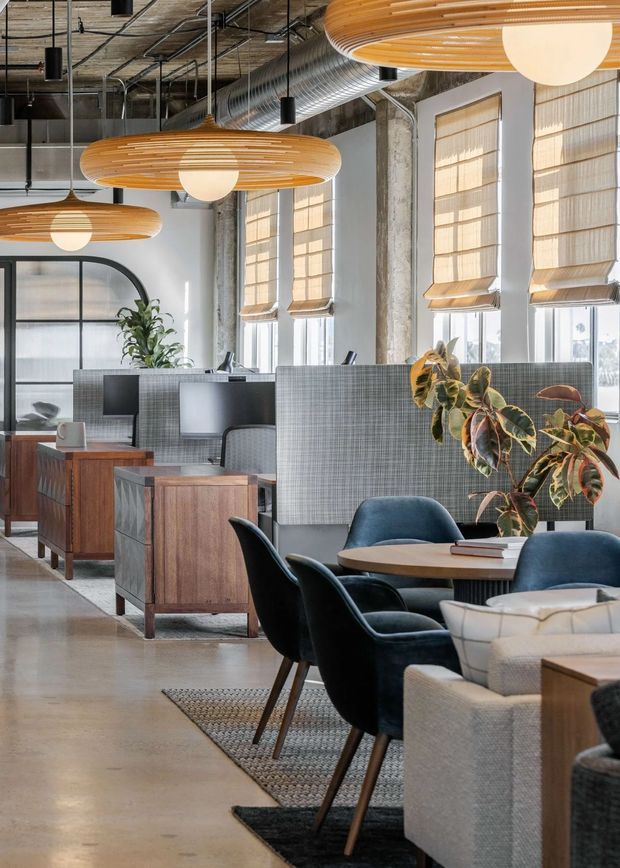A bright, open-plan commercial interior combining a café-style seating area with workstation booths. The space features polished concrete flooring, exposed concrete columns, exposed services and ductwork, large industrial windows with woven roller blinds, grey fabric acoustic partitions, timber storage cabinets, dark upholstered chairs, and distinctive layered timber pendant lights. Indoor plants soften the industrial fit-out.