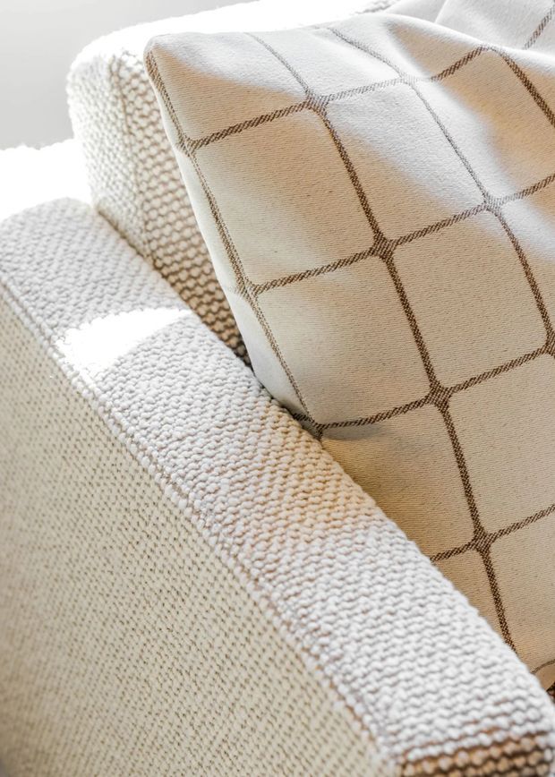 Close-up detail of a softly textured cream upholstered sofa or armchair with a woven boucle-like arm and back, paired with a cream cushion featuring a subtle brown checked grid pattern. The image is brightly lit with a warm, neutral palette and focuses on textile texture and upholstery rather than the wider room.
