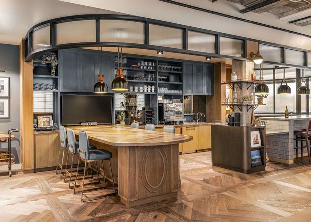 A richly detailed open-plan kitchen and entertaining space with a large curved timber island, integrated dark cabinetry, bar seating, pendant lights, open shelving, and a television. Black-framed glazed partitions with fluted glass create an industrial-style upper screen, while herringbone timber flooring and warm wood finishes add a refined hospitality-inspired character.