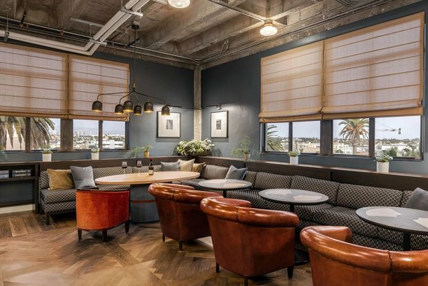 A stylish hospitality-style lounge or dining space with exposed concrete ceilings, visible services, dark painted walls, large windows fitted with woven roller blinds, and herringbone timber flooring. The room contains curved brown leather armchairs, patterned upholstered banquette seating, round dining tables, pendant lighting, framed artwork, and greenery. Palm trees and a waterfront or coastal urban outlook are visible beyond the windows.