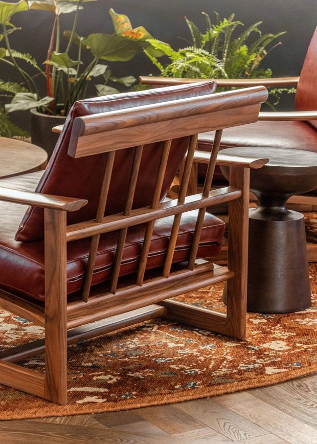 Close-up interior view of a richly styled seating area featuring a sculptural mid-century-inspired wooden lounge chair with vertical slat back, deep reddish-brown leather upholstery, and rounded timber arms. A dark pedestal side table, patterned rust-toned area rug, timber flooring, and lush indoor plants complete the warm, earthy composition.