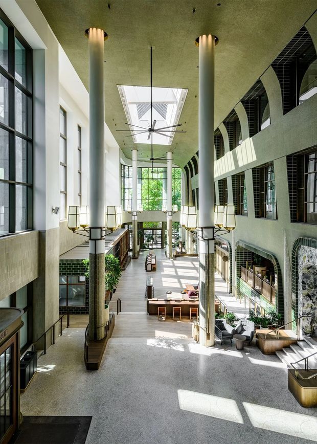 A tall, sunlit multi-level interior atrium or lobby with a dramatic double-height void. Slender cylindrical columns support a textured ceiling with large skylights and oversized ceiling fans. Floor-to-ceiling glazing lines the left side, while the right side features arched openings, dark tiled reveals, and upper-level walkways. Built-in seating, tables, lounge furniture, planters, and layered staircases create a contemporary hospitality or commercial gathering space.