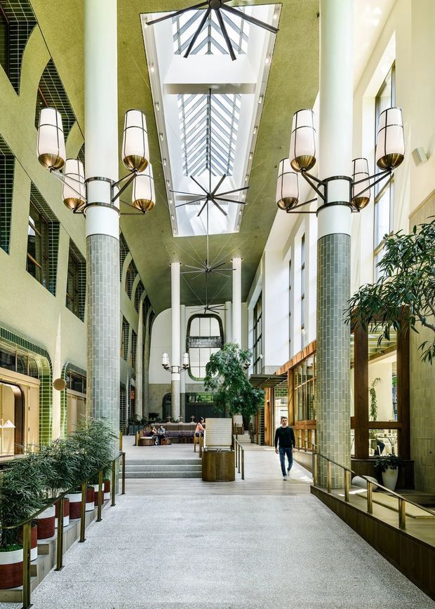 A tall, airy commercial interior atrium or passage with a long skylight and clerestory glazing, exposed geometric ceiling structure, large ceiling fans, decorative multi-arm pendant lights, and slender tiled columns. The space has pale green textured walls, terrazzo-like flooring, timber-framed glazed shopfronts or rooms, integrated seating, and substantial indoor planting. Steps lead toward a lounge area at the far end, with people providing scale.