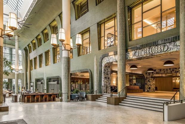 A grand multi-storey commercial hotel or office lobby with a tall glazed atrium, exposed concrete ceiling, large cylindrical tiled columns, patterned masonry walls, arched openings, stepped entry, and warm decorative pendant lighting. Lounge seating, dining tables, and a clearly marked “Tower Lobby” reception entrance are visible.