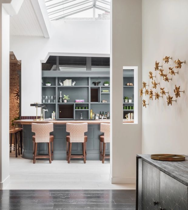 Bright, high-ceilinged contemporary kitchen and home bar area with a large island fitted with patterned upholstered stools, dark grey built-in cabinetry and open shelving displaying glassware and bottles. A roof skylight brings in abundant daylight, while a partial brick wall and dark timber flooring add contrast. The foreground includes a pale stone or patterned built-in console, and a decorative cluster of gold wall sculptures is visible on the adjoining wall.