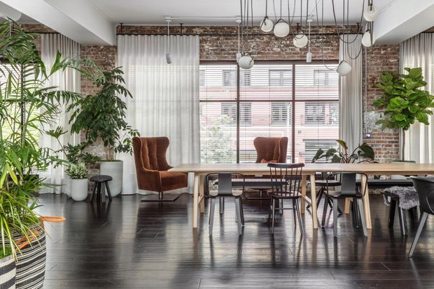A bright open-plan dining and living space with a long timber dining table, mixed dark and light dining chairs, two upholstered lounge chairs, abundant indoor plants, and large industrial-style windows. Exposed red brick walls, dark timber flooring, sheer curtains, white structural beams, and a cluster of suspended pendant lights create a refined industrial interior.