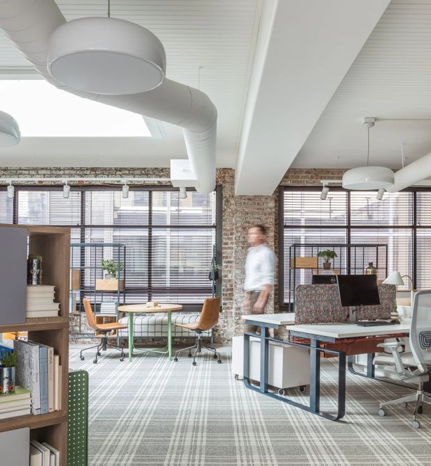 Bright open-plan office interior with exposed brick columns, large industrial-style windows with horizontal blinds, exposed white ductwork, white ceiling beams, and a grey patterned carpet. The space includes shared worktables, rolling office chairs, desks, shelving, plants, and contemporary pendant and task lighting.