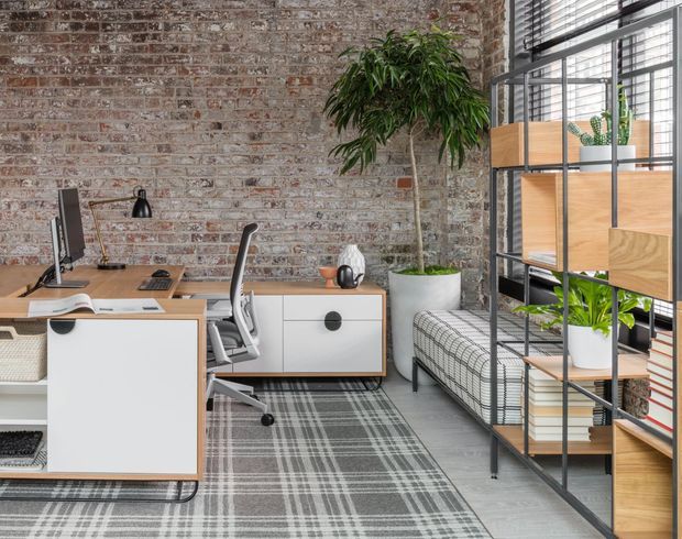 A stylish home office with a contemporary industrial character. The room features exposed brick walls, grey plank flooring, modern white and timber desks with black pulls, a computer workstation, shelving with timber compartments, a patterned grey rug, a mesh office chair, and abundant indoor plants. Black-framed glazing with horizontal blinds brings in natural light, while a cushioned bench sits beside the window.