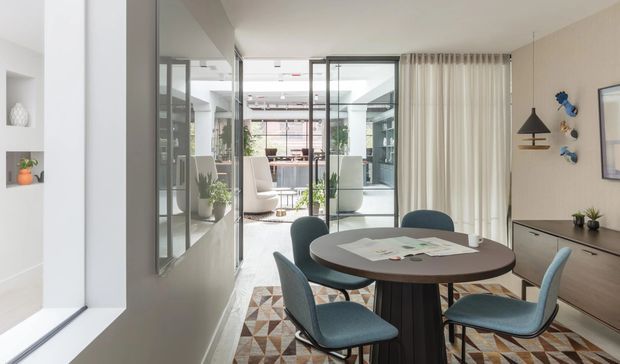 Bright contemporary dining area with a round dark wood dining table and four upholstered blue chairs on a geometric patterned rug. White walls, built-in shelving, a dark timber sideboard, pendant lighting, and full-height glazed sliding doors connect the dining space to an airy open-plan living area beyond. A large interior window and reflected glazing add depth and light.