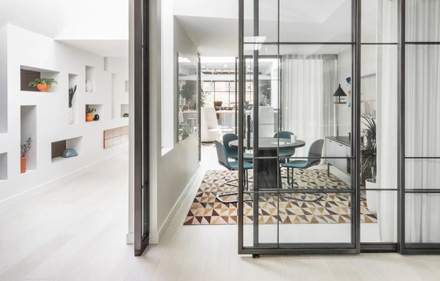 Bright contemporary interior viewed through black metal-framed glazed partitions. The main space is a dining area with a round dark table and upholstered chairs, set over a geometric multicoloured rug. White walls incorporate recessed display niches and built-in joinery, while sheer curtains, indoor plants, and extensive glazing create an airy open-plan feel.