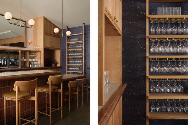 A refined home bar with a dark stone or speckled feature wall, extensive warm timber cabinetry, open shelving for wine glasses, and a polished bar counter with tan upholstered stools. Globe wall and pendant-style lighting, mirrored surfaces, and carefully arranged glassware create an elegant, intimate hospitality atmosphere.