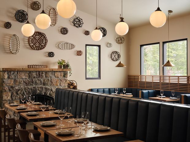 A warmly lit restaurant dining interior with multiple timber tables set with plates and wine glasses, surrounded by dark upholstered banquette seating and dining chairs. A substantial rough stone fireplace with timber mantel dominates the left side, while large windows and a slatted timber screen bring in views of greenery. Numerous sculptural woven wall decorations and suspended globe pendant lights add visual interest.