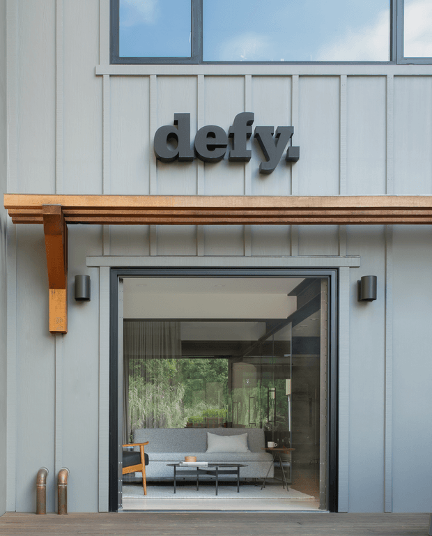 Close-up exterior view of a contemporary commercial or studio-like building entrance. The facade is clad in pale grey vertical panels with dark-framed glazing, a projecting timber-toned canopy supported by timber brackets, wall-mounted cylindrical lights, and a bold dark “deffy” sign. Through the glazed doorway, a modern lounge with a sofa, coffee table, and timber-framed chair is visible.