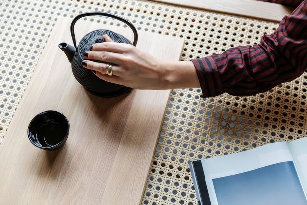 Overhead lifestyle image of a black textured teapot and matching cup placed on a light timber table, with a woven cane or rattan chair surface surrounding it. An open book or magazine is partially visible at the lower right, and a person's hand reaches toward the teapot. The composition has a calm, tactile, contemporary natural-material aesthetic.