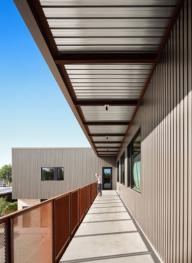 A long elevated exterior walkway or balcony runs alongside a modern building. The facade is clad in narrow vertical ribbed panels, with large dark-framed windows and doors. A translucent corrugated canopy is supported by exposed dark timber or steel beams, while rust-coloured perforated metal balustrades line the open edge. Strong sunlight creates linear shadows across the pale textured walkway, with clear blue sky and limited landscaping visible beyond.