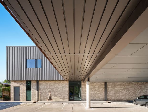 Exterior architectural photograph of a contemporary multi-level building with a deep covered entry or carport-like canopy. The facade combines vertically ribbed grey metal cladding above with pale textured brickwork below, dark-framed glazing and doors, and a broad soffit lined with narrow linear panels. Concrete paving and marked parking bays occupy the foreground.