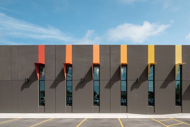 A contemporary commercial building facade viewed straight on, composed of dark grey vertical panels with a repeated rhythm of narrow floor-to-ceiling windows. Each window is framed by a projecting brightly coloured canopy or vertical accent in red, orange, and yellow tones, creating strong geometric shadows across the facade. A paved parking area and clear blue sky are visible.
