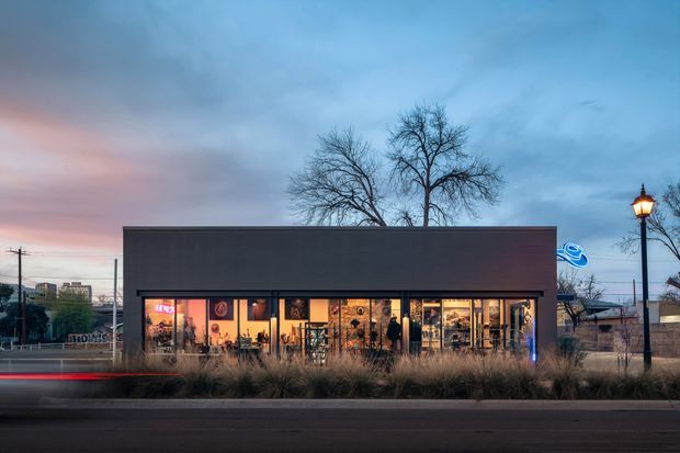 Street-level dusk photograph of a low, rectangular contemporary commercial building with a dark, largely opaque upper facade and a fully glazed storefront along the ground floor. Warm interior lighting reveals a retail or hospitality-style fit-out, while ornamental grasses, a vintage-style street lamp, utility lines, and passing traffic frame the urban streetscape.