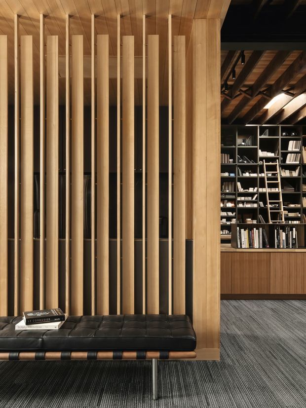 Interior view of a refined library or reading lounge featuring a black tufted leather bench in the foreground, a timber slat screen and warm timber wall panelling, and extensive dark built-in book shelving beneath exposed timber roof rafters. The space has a sophisticated, moody atmosphere with layered architectural joinery and linear carpeting.