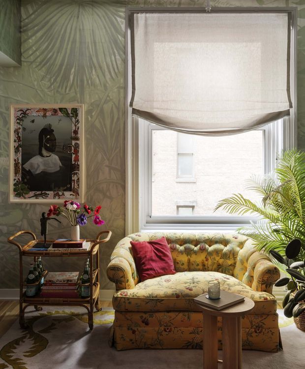 A richly decorated, intimate sitting area with a floral upholstered sofa, red accent cushion, small side table, and a vintage rattan drinks trolley. A large window with a white Roman blind brings in natural light, while botanical wallpaper, framed artwork, flowers, and lush indoor palms create a layered, eclectic atmosphere.