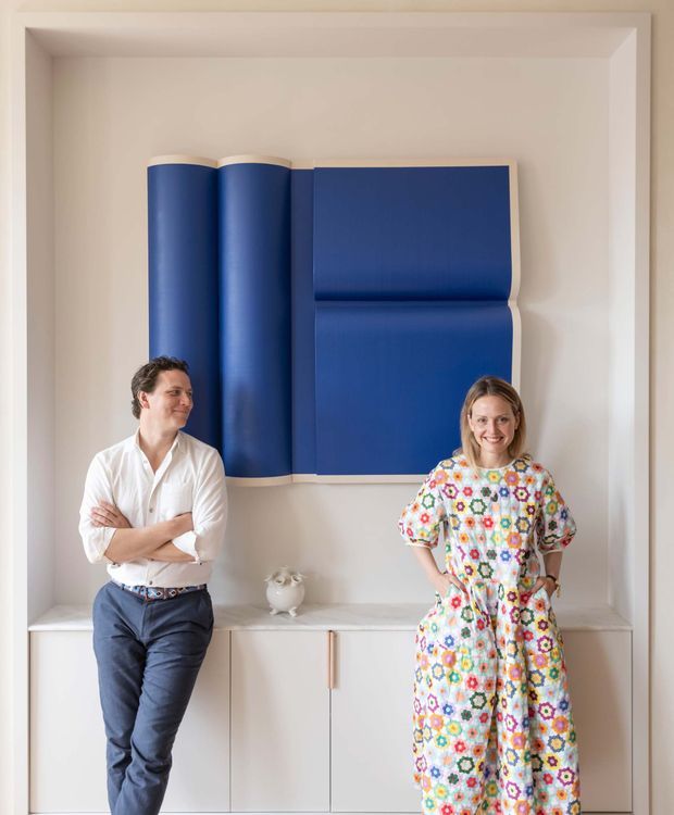 A bright, refined interior vignette with two people standing in front of a built-in pale cabinetry unit and a dramatic sculptural blue wall artwork or upholstered feature. The room has warm off-white walls, a light stone-like benchtop, minimal cabinetry with slim timber-toned pulls, and a recessed architectural wall niche with a framed surround. The styling combines restrained contemporary joinery with a bold, colourful statement piece.