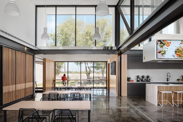 A spacious contemporary open-plan kitchen, dining and living area with a dramatic double-height glazed volume. The room features polished concrete flooring, dark framed floor-to-ceiling glazing, exposed dark structural beams, pendant lighting, a large island kitchen, and timber slatted screening. Dining tables and chairs occupy the foreground, while a landscaped waterfront or park-like view is visible beyond the glazing.