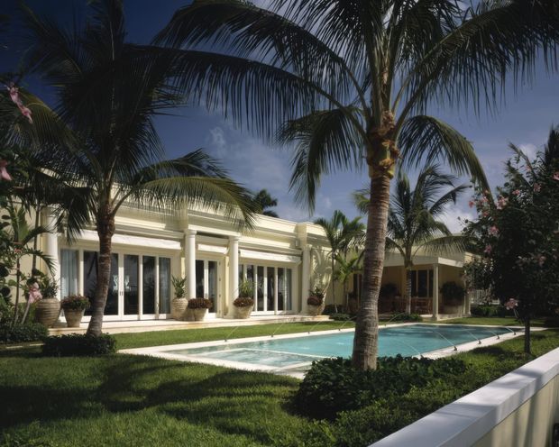A tropical, low-rise villa exterior framed by mature palm trees and manicured lawn, with a long rectangular swimming pool in the foreground. The cream-coloured facade features extensive white-framed glazing, classical columns, deep eaves, and layered horizontal roofline detailing. A low white boundary wall runs along the front edge.