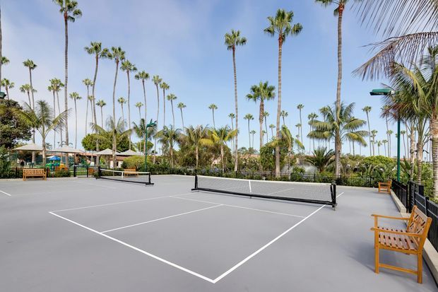 Outdoor pickleball or tennis-style court with a low net, crisp white boundary lines, and perimeter seating. The court is surrounded by lush tropical landscaping, numerous tall palm trees, and resort-like outdoor structures under a bright blue sky.