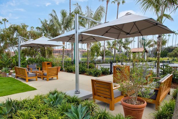 A landscaped outdoor terrace with multiple large shade umbrellas, warm timber lounge seating, concrete paving, lawn, ornamental planting, and palm trees. The space appears designed for relaxed outdoor dining or lounging beside a landscaped recreational area.