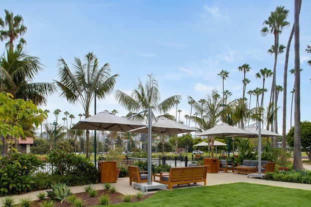 A landscaped outdoor resort-style lounge area with wooden benches and tables beneath large grey umbrellas, surrounded by palm trees, low planting, manicured lawn, and poolside-style fencing. The scene is bright, relaxed, and tropical.