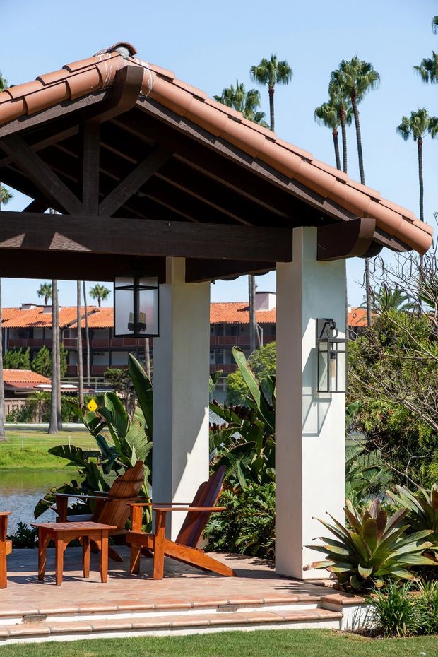 A covered outdoor seating pavilion with white stucco columns, dark exposed timber beams, and a terracotta tiled pitched roof. Wooden lounge chairs and a small table sit on a tiled terrace surrounded by lush tropical landscaping, with palm trees and additional terracotta-roofed buildings visible beyond.