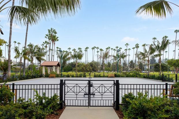 A bright landscaped outdoor entrance with a black metal decorative gate opening onto a paved path. Beyond the gate is a broad paved or court-like area, lush lawns, dense tropical landscaping, numerous palm trees, and a small terracotta-tiled roof pavilion. The scene has a sunny resort-like character and clear views across the grounds.