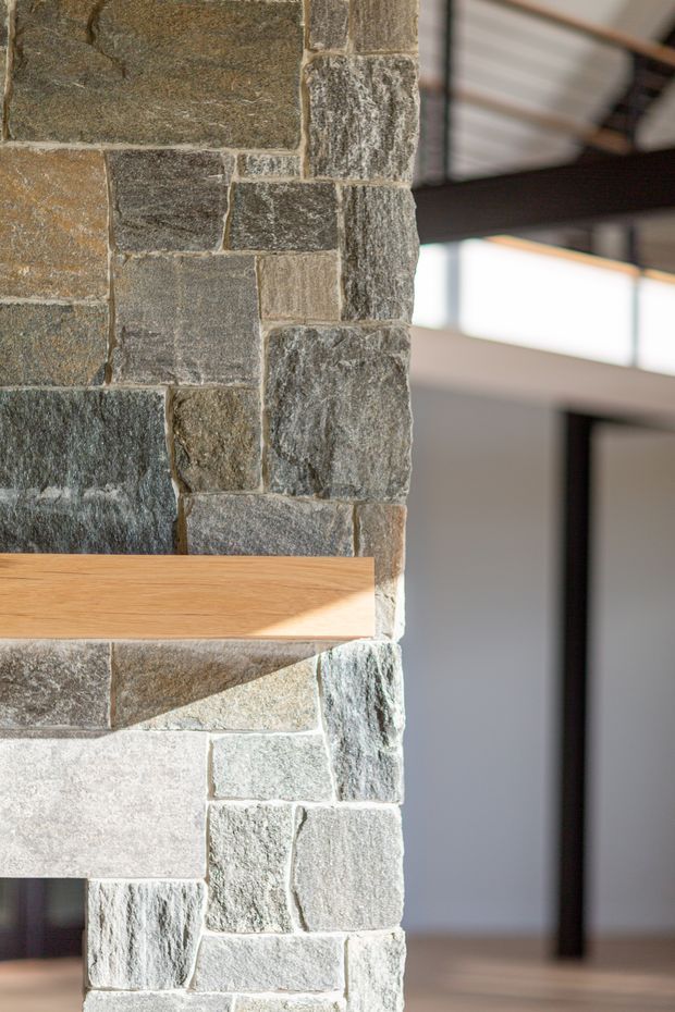 Close-up interior architectural detail showing a rough-textured natural stone masonry wall or chimney with irregular grey, brown and beige stone blocks. A simple timber shelf projects from the wall, with a contemporary interior and exposed dark structural beam visible in the background.