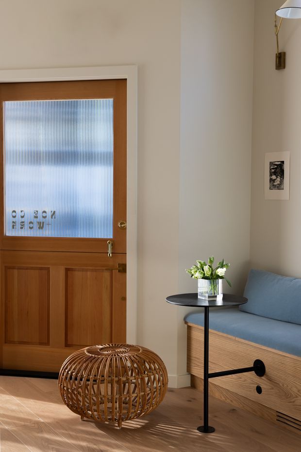A warmly lit residential entryway with a timber panelled door featuring a ribbed translucent glass insert, opening onto a neutral plastered interior. The space includes pale timber flooring, a built-in timber bench with a blue upholstered cushion, a slim black side table with a glass vase of flowers, a sculptural woven rattan ottoman, a wall-mounted brass reading light, and a small framed artwork. The composition has a refined, contemporary yet warm character.