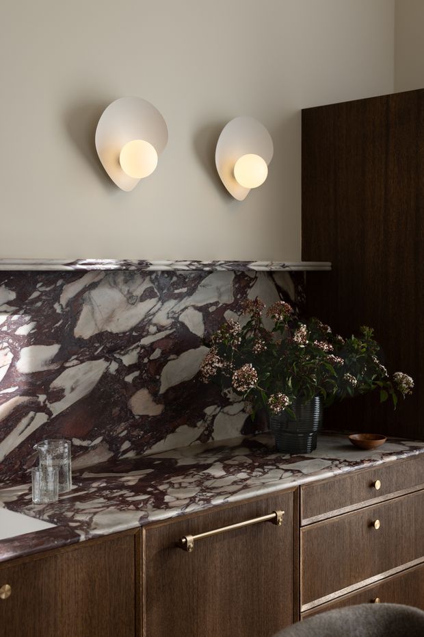 A refined interior vignette featuring a dark timber sideboard or built-in cabinet with brass hardware, topped by a dramatic veined natural stone surface and matching stone backsplash. Two sculptural wall sconces cast warm light onto a neutral plaster wall, while a vase of foliage and small decorative objects add softness. The composition is luxurious, moody, and highly material-focused.