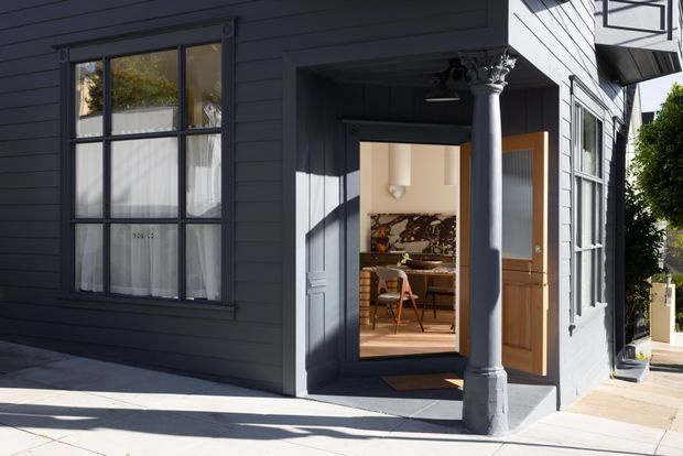Street-facing exterior of a dark blue-grey weatherboard house with tall multi-pane windows, a recessed entry porch, and a substantial classical-style column. Through the open doorway, a warmly lit kitchen or dining area with timber cabinetry and a patterned backsplash is visible.