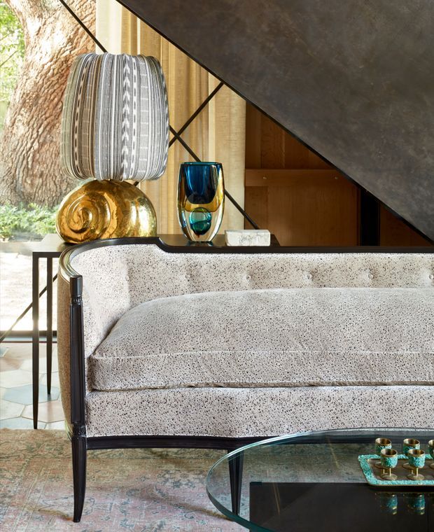 Close-up interior view of a sophisticated living room, centered on a textured pale upholstered sofa with a dark slim frame. A round glass coffee table, patterned area rug, sculptural decorative objects, and a large striped pendant light create a refined, eclectic setting. Timber cabinetry, expansive glazing, and a tree visible outside form the background, while a dark angular architectural element crosses the upper right of the image.